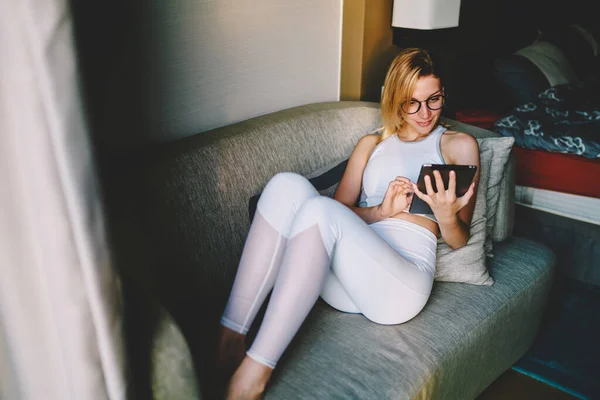 身穿白色运动服 戴着眼镜 面带微笑的金发年轻女子躺在舒适的沙发上 房间里放着靠垫和浏览平板电脑 — 图库照片