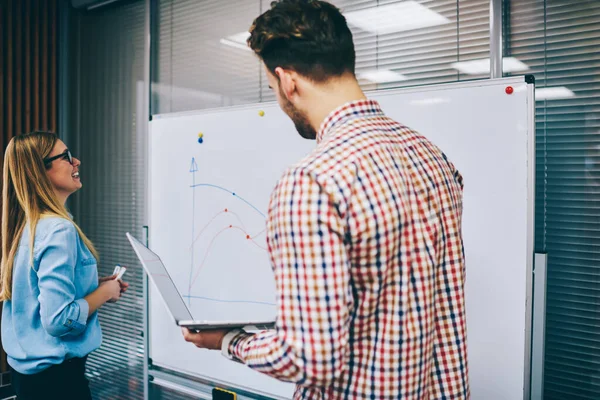 Gündelik giysiler içinde neşeli genç iş arkadaşları ofiste yeni bir projede çalışırken dizüstü bilgisayar kullanarak büyük beyaz tahtada grafikler çiziyorlar.