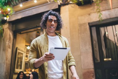 Şehirde terasta durup tableti elinde tutan, gündelik giyinen düşünceli, etnik bir erkek.
