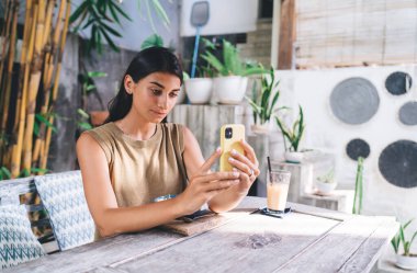 Yazlık kıyafetli iyimser kadın, rahat terasta masada otururken akıllı telefon kullanıyor ve öğle yemeği molasında ferahlatıcı smoothie içiyor.