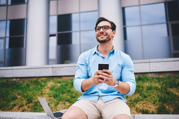 Smiling handsome student laughing receiving funny video from friend in social networks recreating outdoors, hipster guy enjoying 4G connection in university campus sending photos and video on mobile