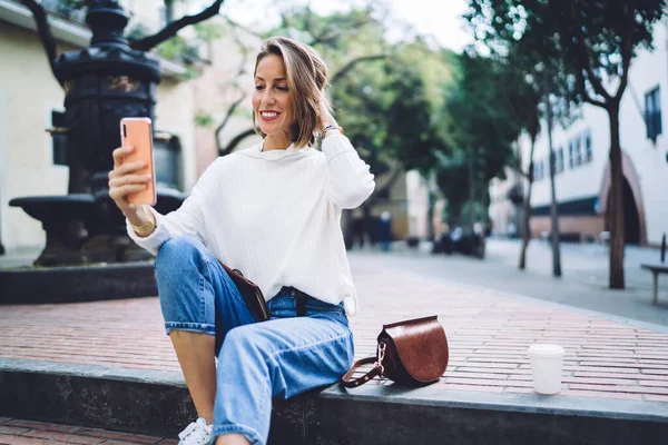 Positive female tourist in trendy outfit taking self portrait on mobile phone while relaxing on stairs of park against fountain after sightseeing