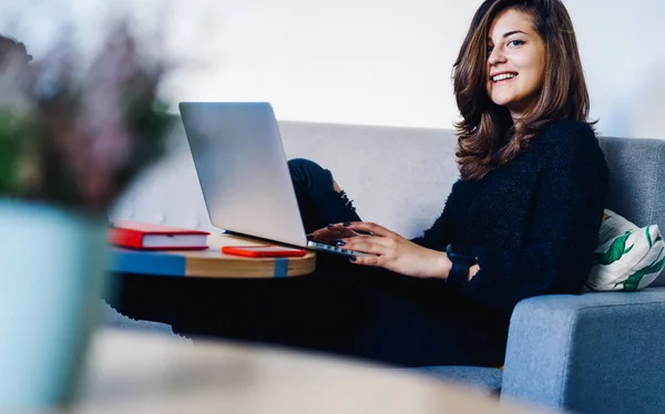 現代のカフェテリアの快適なソファにノートとスマートフォンでテーブルに座っている間 コンピュータ上でリモートで働く笑顔の若い女性 — ストック写真