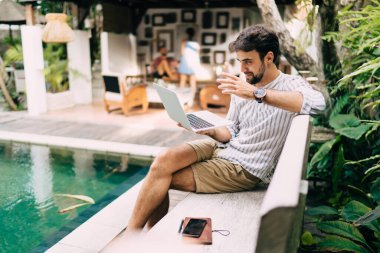 Yan bakış, şık yaz kıyafetleri içinde genç bir erkeğin yüzme havuzunun yanında bankta oturması ve tropikal tatil beldesinde netbook 'a göz atması.