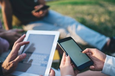 İsimsiz arkadaşların ekranlara dokunması, akıllı telefon ve tabletle mesajlaşması ve parkta çimenlerde yan yana oturması.