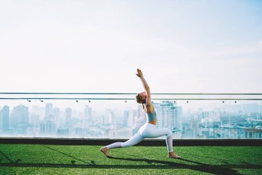 Spor yaparken ince kadın kıyafetlerini giyer, yoga yapar ve güneşli yaz gününde yeşil çatıda Virabhadrasana pozu verir.