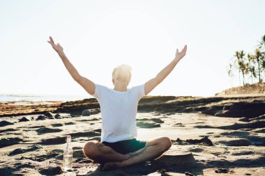 Arkadaki tanınmayan adam, yaz kıyafetleri içinde kumlu sahilde poz veriyor ve kollarını güneşe kaldırıyor.