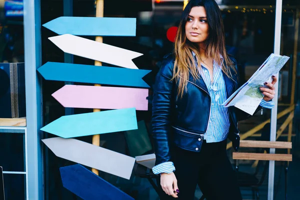 Crop pensive female in casual clothes with long hair looking away standing on street near signs and orientating in city using map