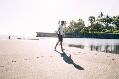 Genç sakallı, spor kıyafetli genç adam okyanus yakınlarındaki tropikal bir yerde sabah yürüyüşü yaparken kumlu sahilde yürüyor.