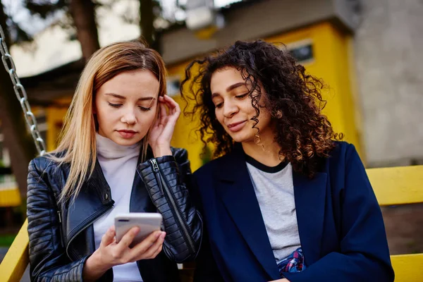 Gündüz vakti sokakta bankta oturan ve arka planda bina inşa ederek akıllı telefonlarda sörf yapan çok ırklı genç arkadaşlar.