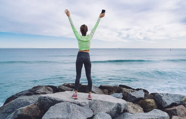  Kulaklık takmış, müzik dinleyen bir kadın. Kıyı şeridi taşlarında el kaldırırken.