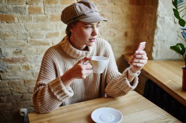 Yukarıdan, şık giyinmiş, cam kenarındaki masada oturan, elinde bir fincan sıcak içecekle video çağrısı yapan genç bir kız öğrenciden.