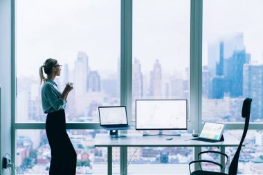 Resmi kıyafetler içinde zarif bir kadın kahve molası veriyor ve aletlerle çalıştığı ofisten şehir manzarasına bakıyor.