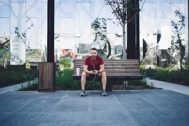 Spor kıyafetleri içinde, elinde plastik bir şişe su, elinde akıllı telefon, tahta bir bankta oturmuş müzik dinleyen odaklanmış bir erkek sporcu.