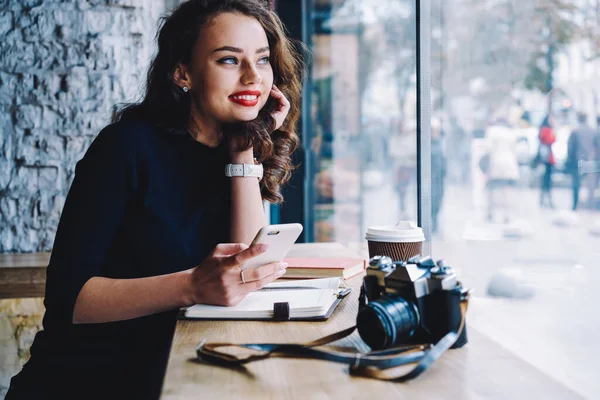 在当代的工作空间里 红唇的快乐女性摄影师拿着智能手机坐在桌子边 一边看一边看 — 图库照片
