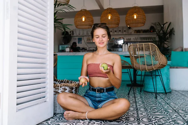 Full body content female in casual clothes smiling and holding apples in hands while sitting on floor in lotus pose on Bali