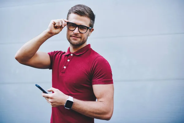 Calm male with beard in trendy outfit and wristwatch adjusting eyeglasses and looking at camera against gray wall of building in daytime