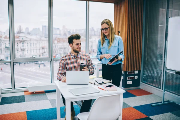 現代のオフィスでノートパソコンやノートブックとテーブルに座っている男性同僚にクリップボードを使用して新しい創造的なアイデアを実証眼鏡で笑顔女性マネージャー — ストック写真