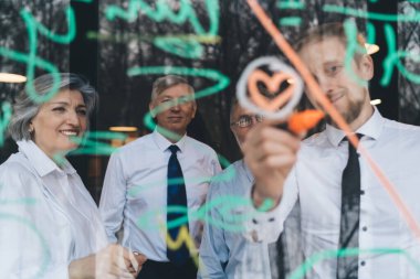 Birlikte iş projesinde çalışırken resmi kıyafetler içinde şeffaf tahtaya notlar yazan iş ortaklarının camından.