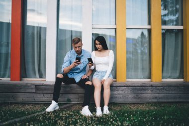 Ciddi, çok kültürlü bir çift ahşap bankta otururken ve parkta birlikte takılırken akıllı telefonları karıştırırken gündelik kıyafetlerini giyiyor.