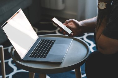 Tanımlanamayan siyah gömlekli, kol saati olan bir erkek oturma odasında kahve masasının yanında oturuyor ve dizüstü bilgisayarla çalışırken cep telefonu kullanıyor.