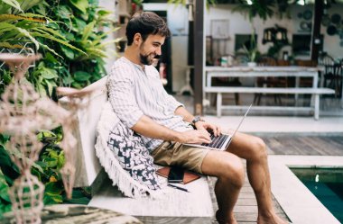 Yazın gülümseyen pozitifler, modern netbook klavyesinde yazarken egzotik tatil beldesindeki havuz kenarında rahat bir bankta takılıyor.