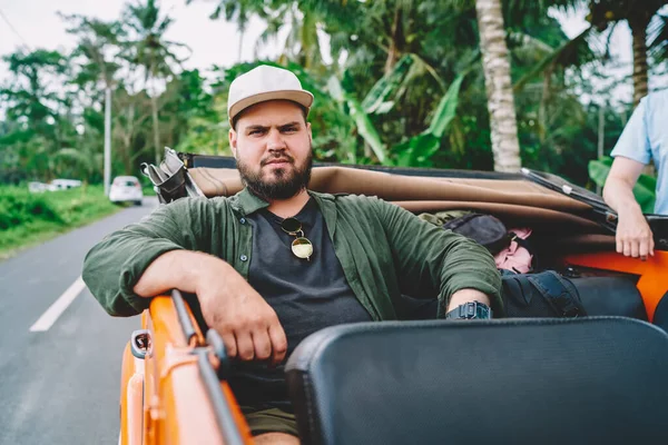 Gloomy man with beard in cap looking at camera and sitting in car on road near tropical forest on summer day