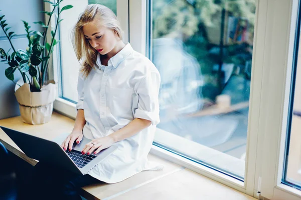 窓辺に座って画面を見ながらノートパソコンを使って新しいプロジェクトに取り組むカジュアルな白い服の若い女性に焦点を当て — ストック写真