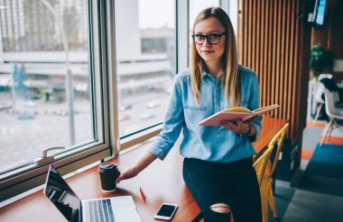 Zeki, sıradan giyinmiş, gözlüklü, elinde kağıt bardak sıcak içecek ve kitapla masada dizüstü bilgisayarlı ve akıllı telefonlu genç sarışın bir kadın.
