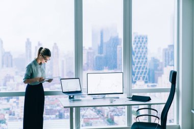 Modern ofisin penceresinin yanında duran ve çalışırken tablet ekranına yoğunlaşan resmi giysili ciddi kadın gözlüğü.