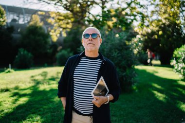 Modern dijital tablet taşıyan ve güneşli halk parkında poz veren şık güneş gözlüğü takan modaya uygun giyinmiş emekli.