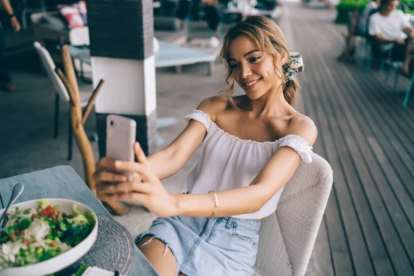 Young female wearing white blouse and denim skirt while sitting at restaurant veranda with salad dish and taking photo on smartphone