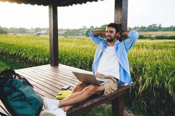 Young smiling bearded remote employee with netbook and hands behind head sitting with crossed legs in pavilion while looking away in summer