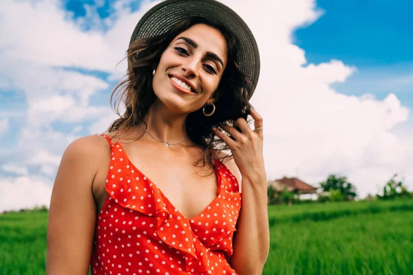 Positive female in dress and hat standing in green field in Bali in sunny summer day and looking at camera
