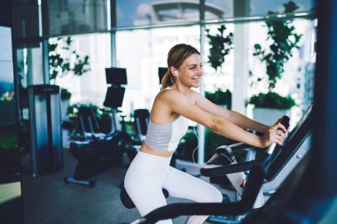 Hafif ferah spor salonunda spor yaparken spor bisikletinin üzerinde oturan dişlek tebessümlü kadın yan görünüşü