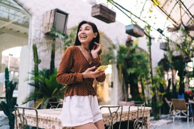 Akıllı telefon kullanırken ve kafede zaman yolculuğu planlarken gülümseyen ve başka yöne bakan içerikli bir kadından.