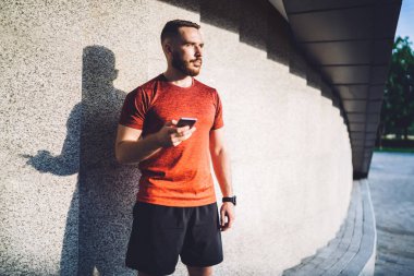 Genç sakallı sportif giysiler giyen erkek sporcu güneşli bir günde binanın karşısında taş duvarla dikilirken cep telefonunu karıştırıyor.