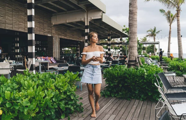Full body of young barefoot female in casual outfit with bare shoulders strolling on wooden terrace of tropical resort while messaging on mobile phone