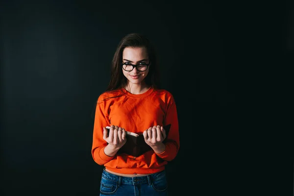 Smiling ark haired woman in orange sweater and jeans looking at camera while standing with book in hands on dark background
