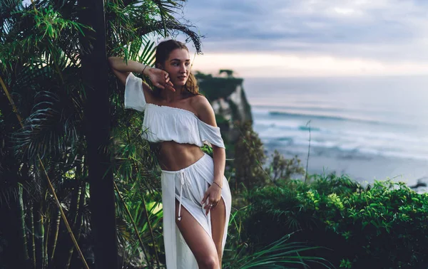 Charming young skinny woman in white elegant two piece dress leaning against poll and looking away with dreamy smile while resting near ocean