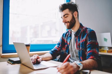 Başarılı sakallı öğrenci web sitesinde dizüstü bilgisayardan web sayfası okuyarak ve not defterine not alarak üniversitede masada oturuyor. Pozitif hippi çocuk ödevleri için bilgi okuyor.