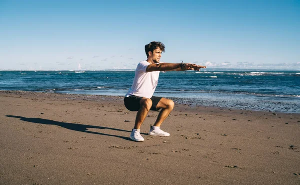 Full body of active bearded male in sportswear and sneakers doing squats on sandy beach near seashore during fitness workout on clear summer day