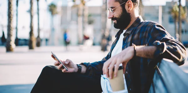 Bearded blogger watching mobile video vlog during leisure time in city resting on bench