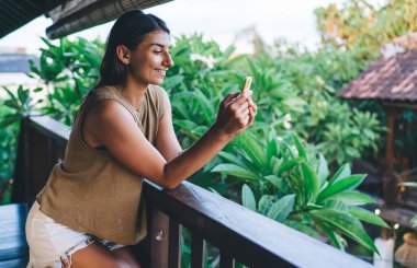 Terasta, egzotik bitkilerin yanında ahşap çitlerin yanında dikilen neşeli etnik kadın manzarası tropik tatil beldesinde cep telefonundan mesaj yazarken...