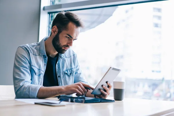 Focused bearded entrepreneur in casual clothes sitting at table with documents and browsing tablet while working on project in contemporary workspace in daylight