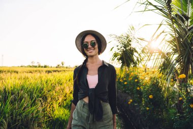 Güneş gözlüklü ve hasır şapkalı neşeli bir kadın turist güneşli bir günde kırsalda yürürken Vietnam 'da tek başına seyahat etmenin keyfini çıkarıyor.