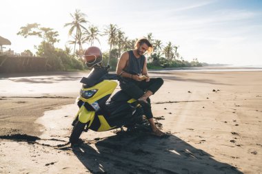Full length of brutal male biker sitting on motorcycle and messaging via smartphone while spending time on beach near sea