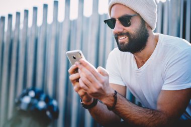 Moda güneş gözlüklü ve kış şapkalı Orta Doğulu bir erkek bankacılık uygulaması ve hesap detaylarını kontrol etmek için akıllı telefon aygıtı kullanıyor.