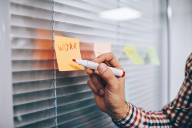 Pencerenin yanındaki parlak ofis odasında duran ve renkli yapışkan kağıda kırmızı kalemle mesaj yazan isimsiz bir adam.