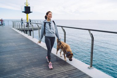 Neşeli, spor elbiseli, spor ayakkabılı, tasmalı genç bir kadın tasmalı bir köpek ve deniz manzarasının tadını çıkarıyor.